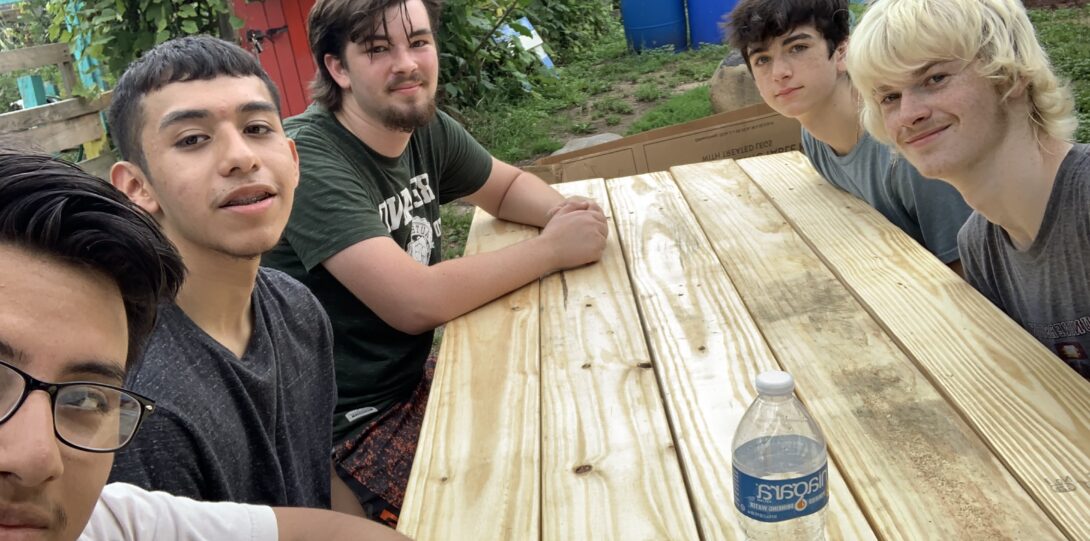 Interns, alonside undergraduate intern Freddy Hernandez, resting on wooden bench they built volunteering at Roots and Rays Community garden